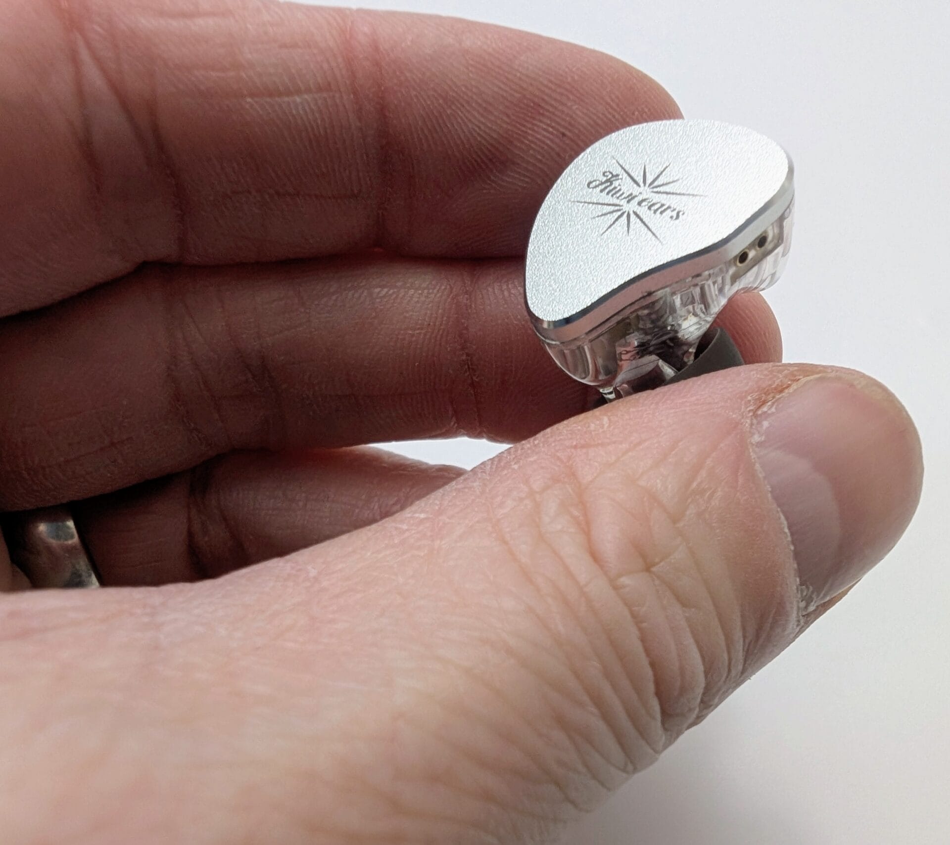 A human hand holding a single Kiwi Ears Belle in-ear monitor (IEM). The silver metallic faceplate with the engraved Kiwi Ears starburst logo is clearly visible, along with the transparent shell showing the internal driver and two-pin connector. The earpiece is held between the thumb and fingers against a plain white background.