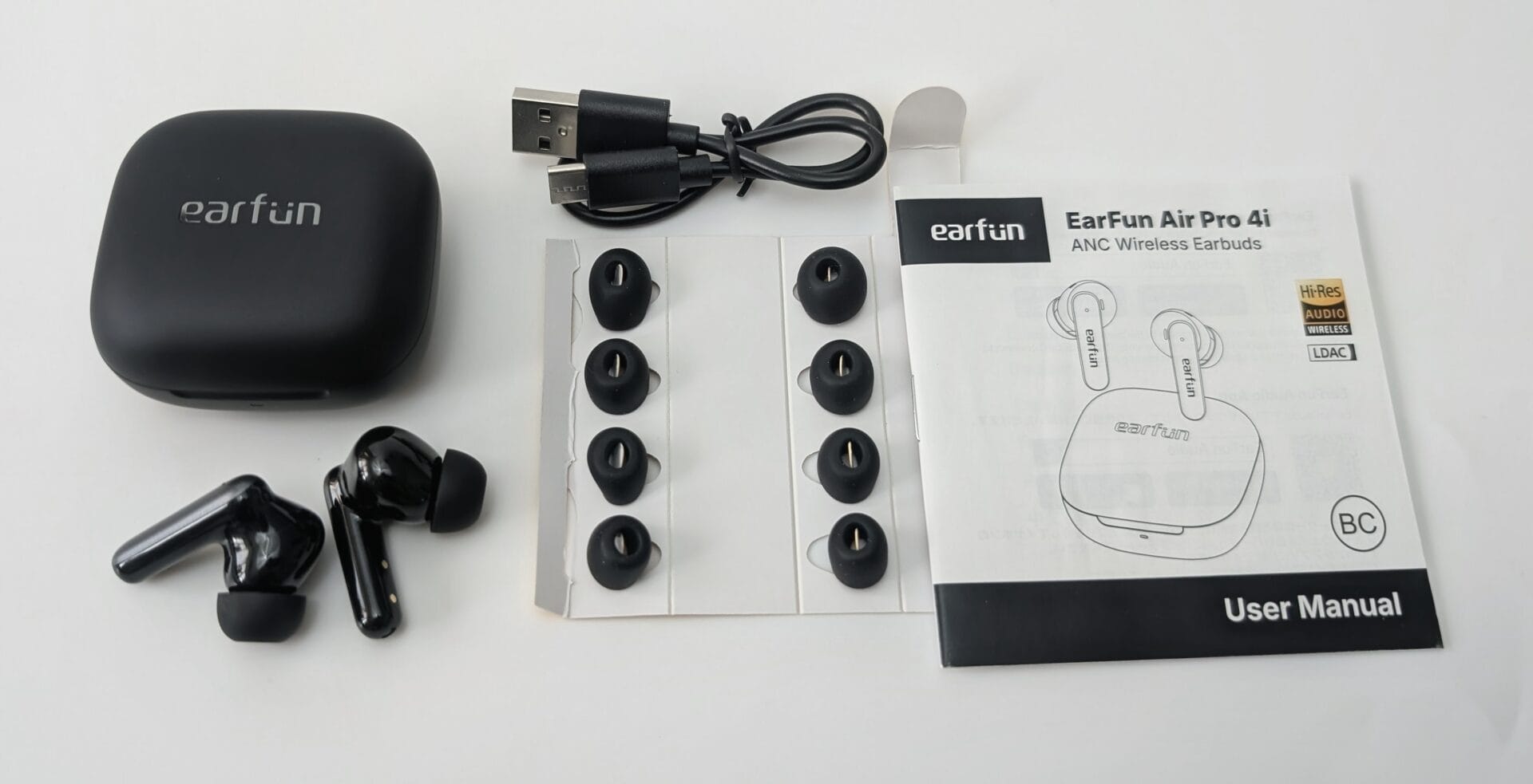 Flat lay of EarFun Air Pro 4i ANC Wireless Earbuds contents on a white background. Shown are: a black charging case with the earfun logo, two glossy black wireless earbuds, a coiled USB-C charging cable, multiple sets of black silicone ear tips in various sizes on a white tray, and the official User Manual booklet displaying the product name, diagrams, and Hi-Res Audio / LDAC logos.