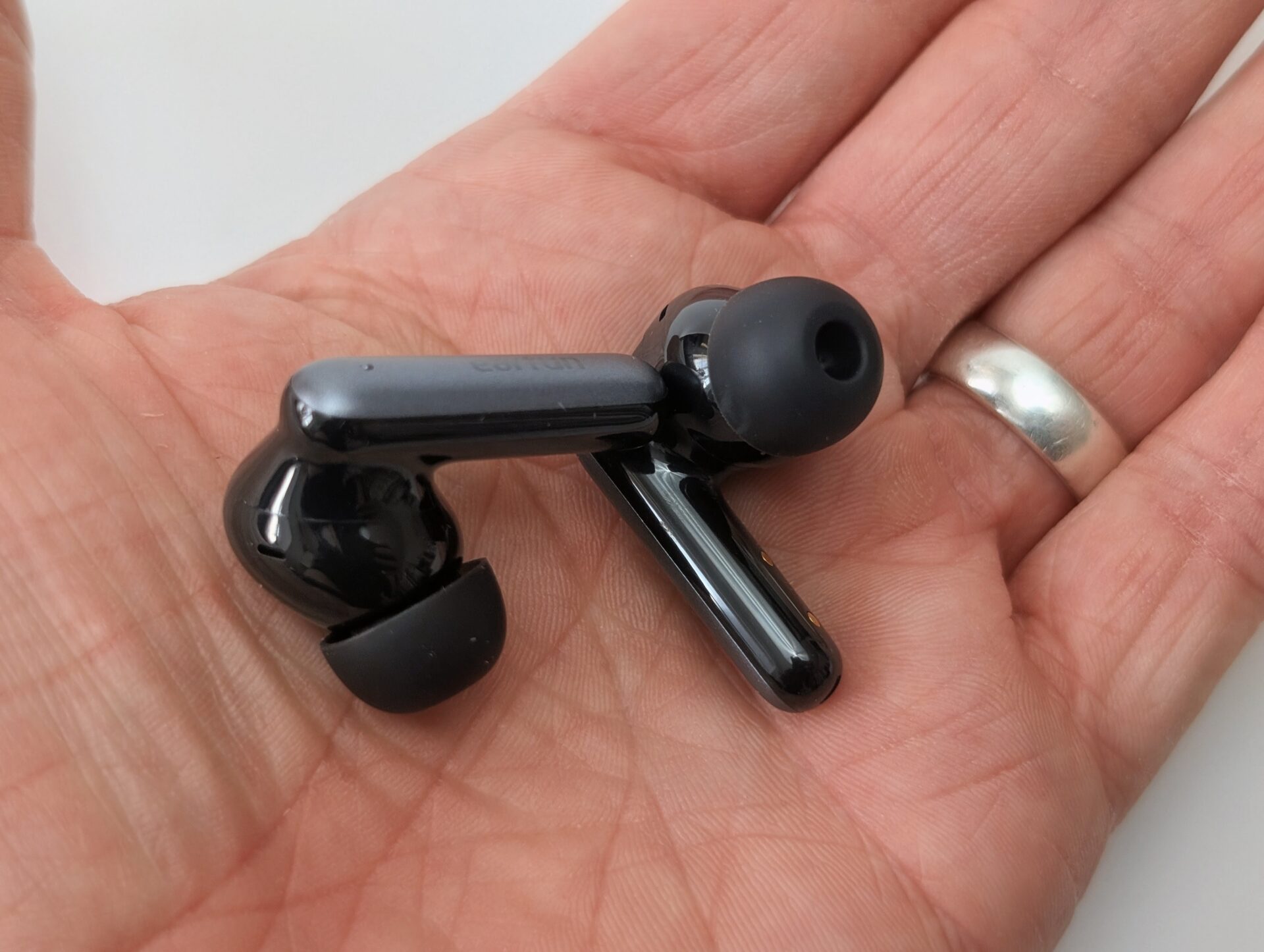 Close-up of a pair of glossy black EarFun Air Pro 4i wireless earbuds resting in the palm of a person's hand. The earbuds feature black silicone ear tips, sleek stems with the 'earfun' logo visible on one, and a premium shiny finish. A silver ring is visible on the person's finger.