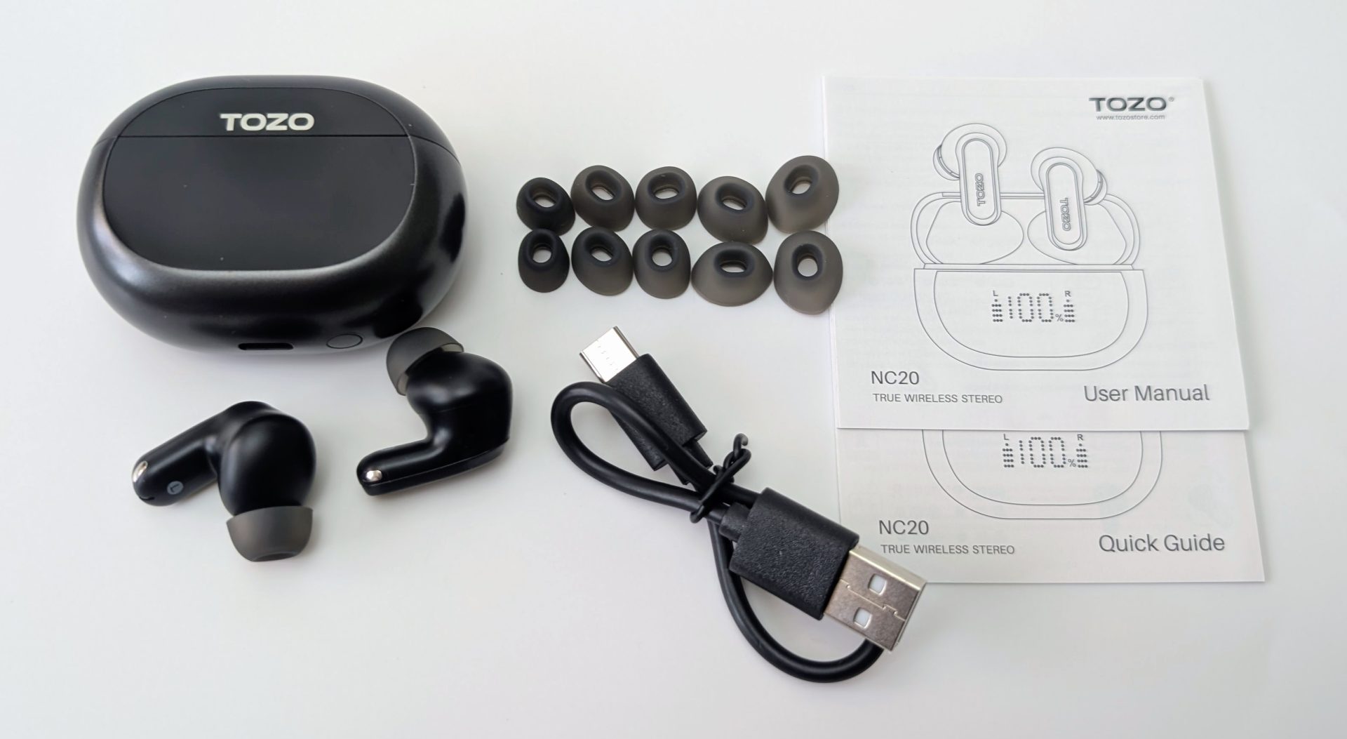 TOZO NC20 Earbuds Flat lay product unboxing of TOZO NC20 true wireless earbuds on a white surface. Items include: a closed glossy black charging case with TOZO logo, two black earbuds placed nearby, multiple pairs of replacement silicone ear tips, a black USB-C charging cable, and TOZO user manual and quick guide booklets.