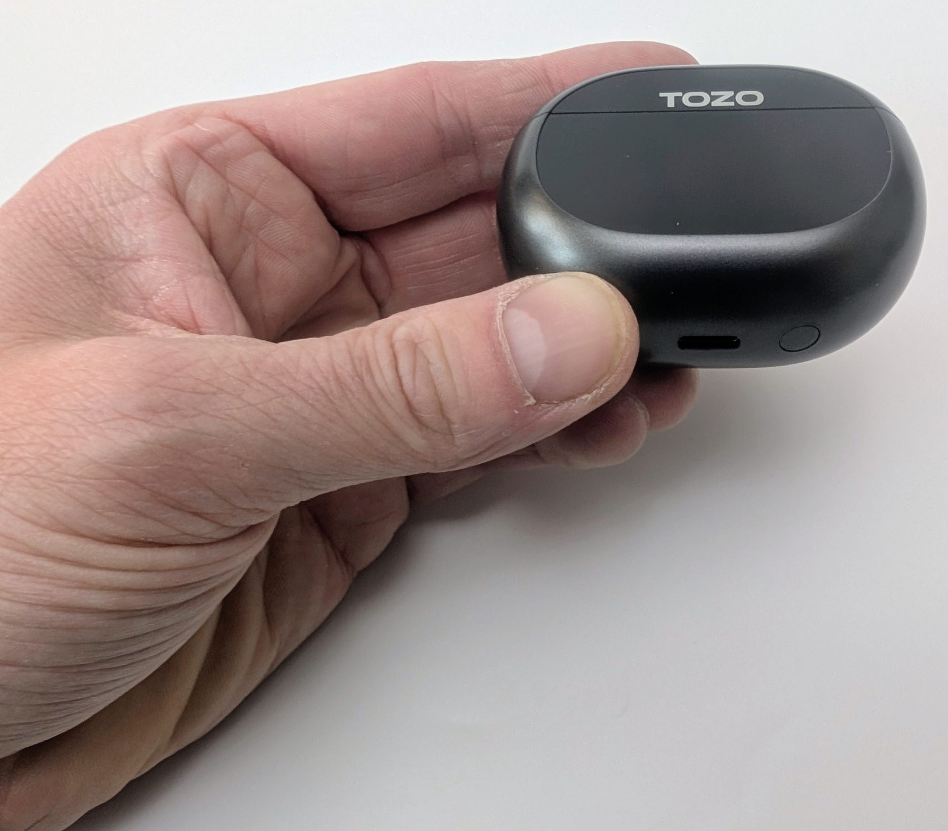 TOZO NC20 Earbuds Human hand holding the closed black TOZO NC20 charging case, showing the glossy top with the white 'TOZO' logo, USB-C charging port, and indicator button on the side.
