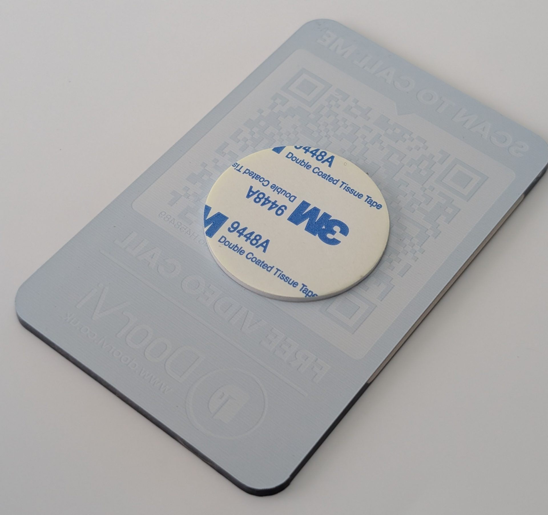 A light gray DoorVi adhesive card shown from the back. A round white 3M 9448A Double Coated Tissue Tape sticker is centered on the surface. Faint embossed text is visible underneath, including 'SCAN TO CALL ME', 'FREE VIDEO CALL', the DoorVi logo, and the website www.doorvi.co.uk.