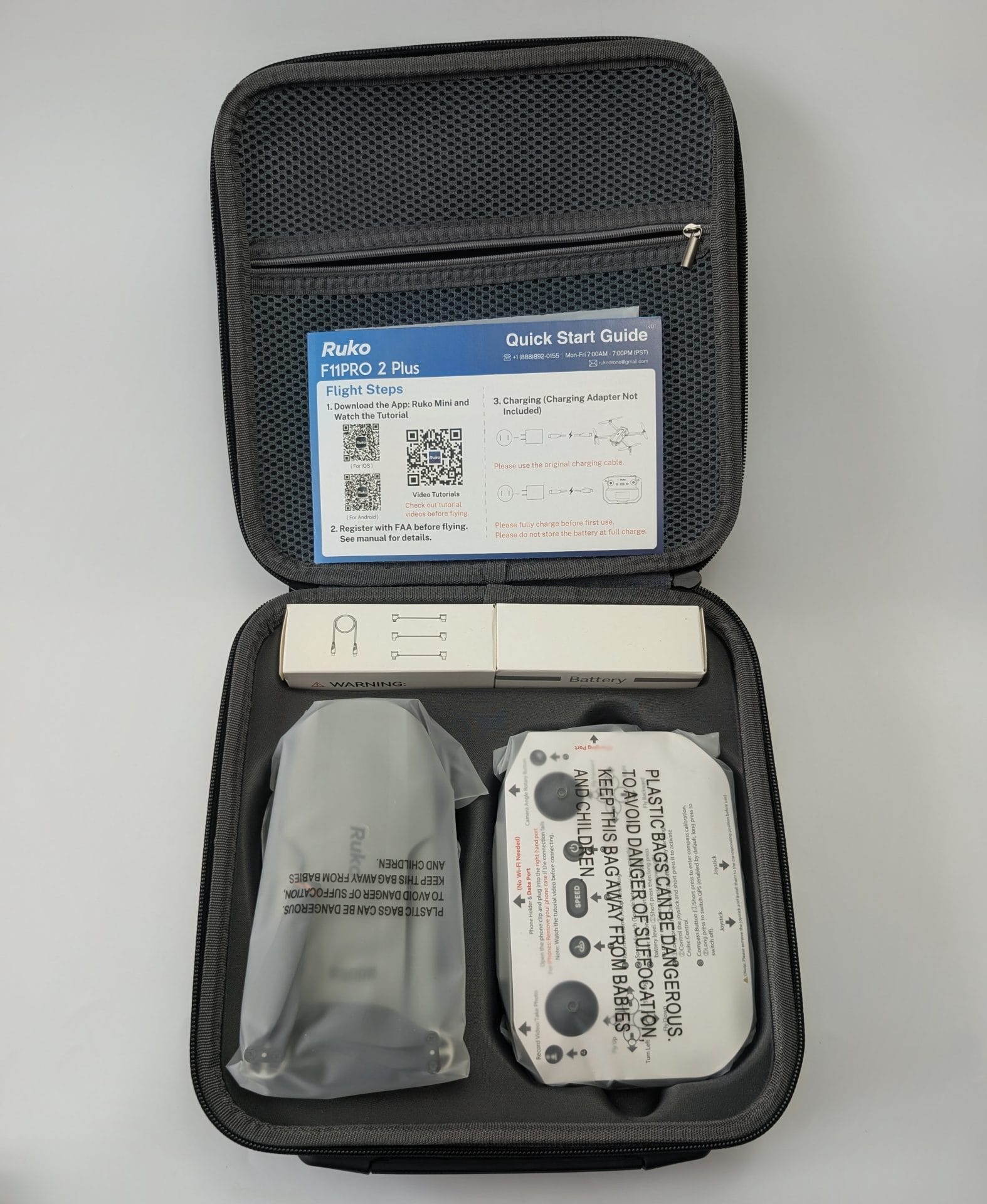 Open black carrying case for the Ruko F11PRO 2 Plus drone, displaying its contents: a Quick Start Guide with flight instructions and QR codes at the top, a white battery pack and cable box in the upper compartment, the drone wrapped in a protective plastic bag on the left, and the remote controller in its packaging on the right.