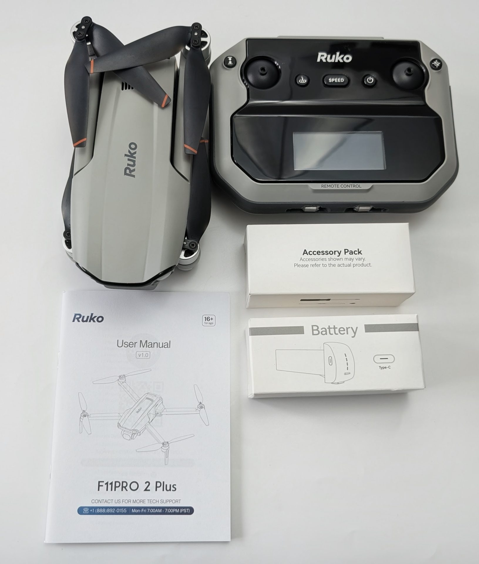 Flat lay of the Ruko F11PRO 2 Plus drone kit on a white background, showing the folded gray drone with black and orange-tipped propellers on the left, the black and gray remote controller with screen and buttons on the right, and below them the User Manual, Accessory Pack box, and Battery box neatly arranged.