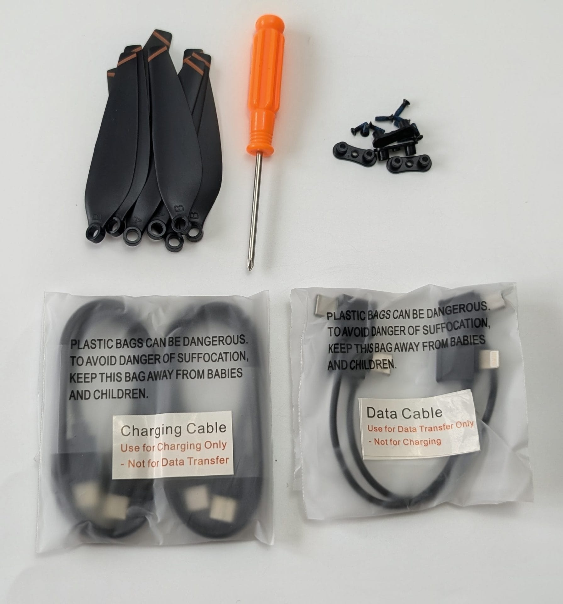 Accessories for the Ruko F11PRO 2 Plus drone laid out on a white surface: a set of spare black propeller blades with orange accents (top left), an orange-handled screwdriver (center), small black screws and propeller guards (top right), a charging cable in a sealed plastic bag (bottom left), and a data cable in a sealed plastic bag (bottom right).