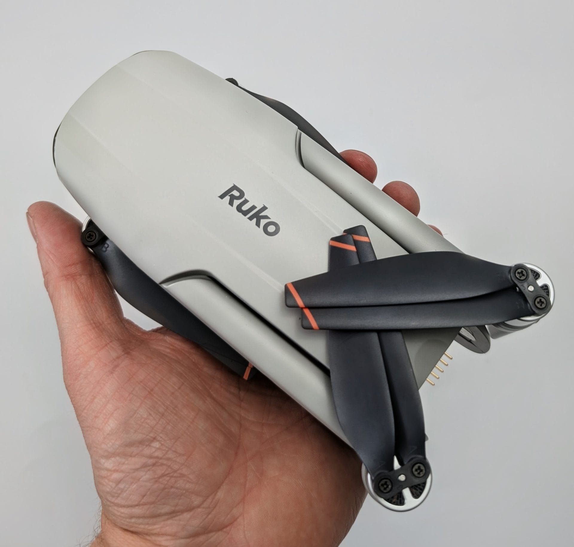 A human hand holding the folded Ruko F11PRO 2 Plus drone against a plain white background, showcasing its compact gray body with the 'Ruko' logo, black propellers featuring orange accents, and visible gold connector pins.