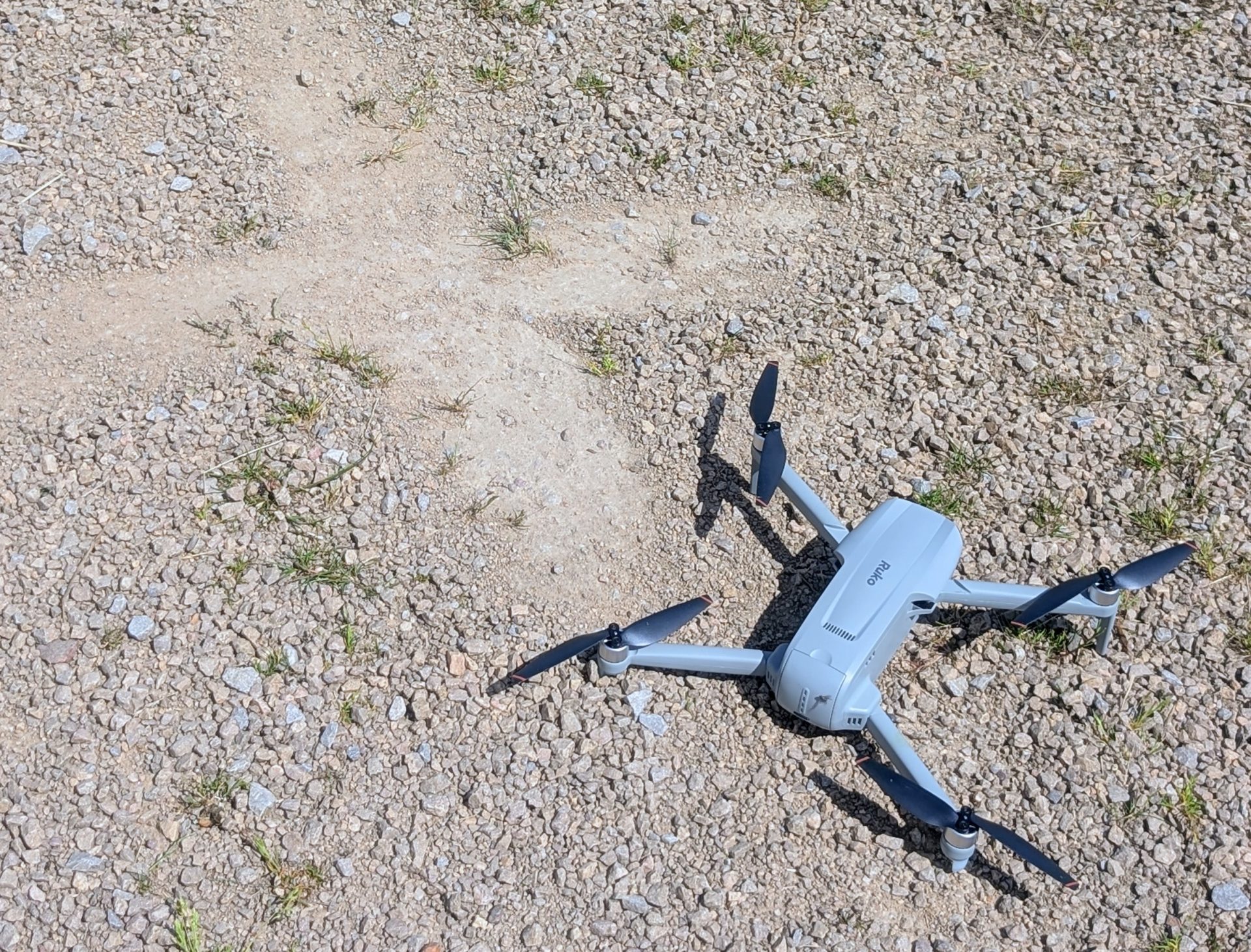 The Ruko F11PRO 2 Plus drone fully unfolded and sitting on a sunny gravel path, with all four black propellers extended and its gray body visible from a high angle.