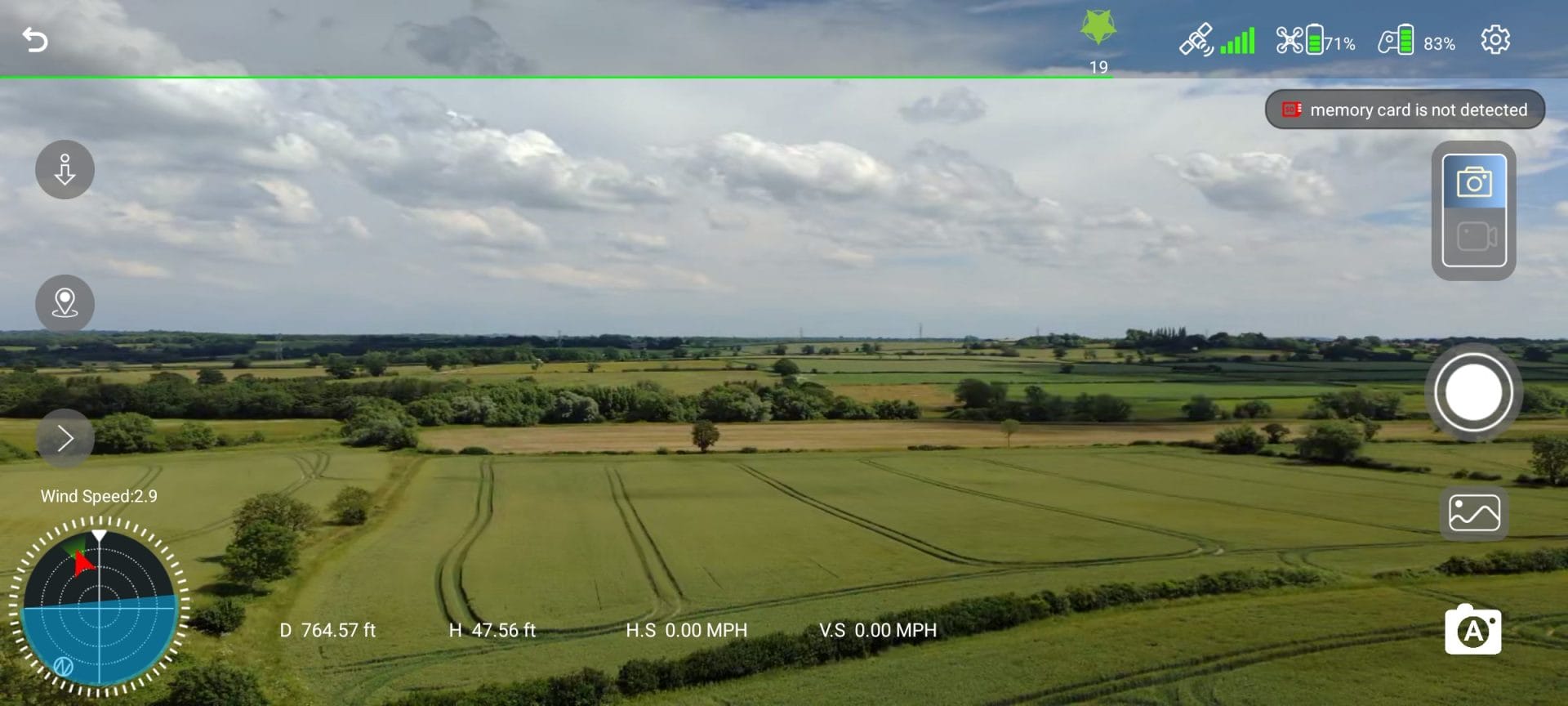 Screenshot of the Ruko F11PRO 2 Plus drone’s live camera feed showing an aerial view of vast green farmland and countryside under a partly cloudy sky. The drone’s flight interface overlay displays telemetry data including 764.57 ft distance, 47.56 ft height, wind speed 2.9, battery levels, and a prominent warning: 'memory card is not detected'.