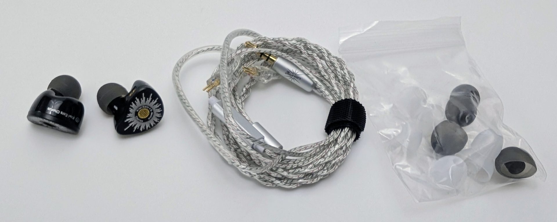 Kiwi Ears Chorus Contents of the Kiwi Ears Chorus IEM set laid out on a white surface: a pair of glossy black in-ear monitors (one showing the outer shell with “Kiwi Ears Chorus” text, the other showing the circular faceplate with white radial pattern and gold center mesh), a coiled silver braided cable with gold 2-pin connectors, silver Y-split and 3.5 mm plug secured by a black Velcro strap, and a clear plastic bag containing assorted silicone ear tips in various sizes and colors.