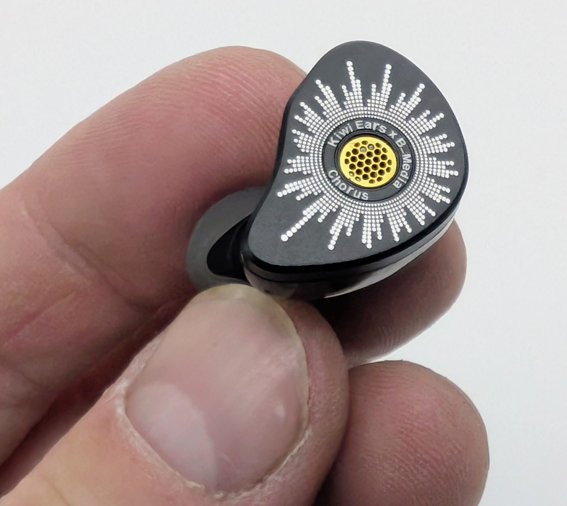 Kiwi Ears Chorus Close-up of a glossy black Kiwi Ears x B_Media Chorus in-ear monitor held between fingers, showing the circular faceplate with a white radial dotted pattern surrounding a central gold mesh grille and the text “Kiwi Ears x B_Media Chorus.”