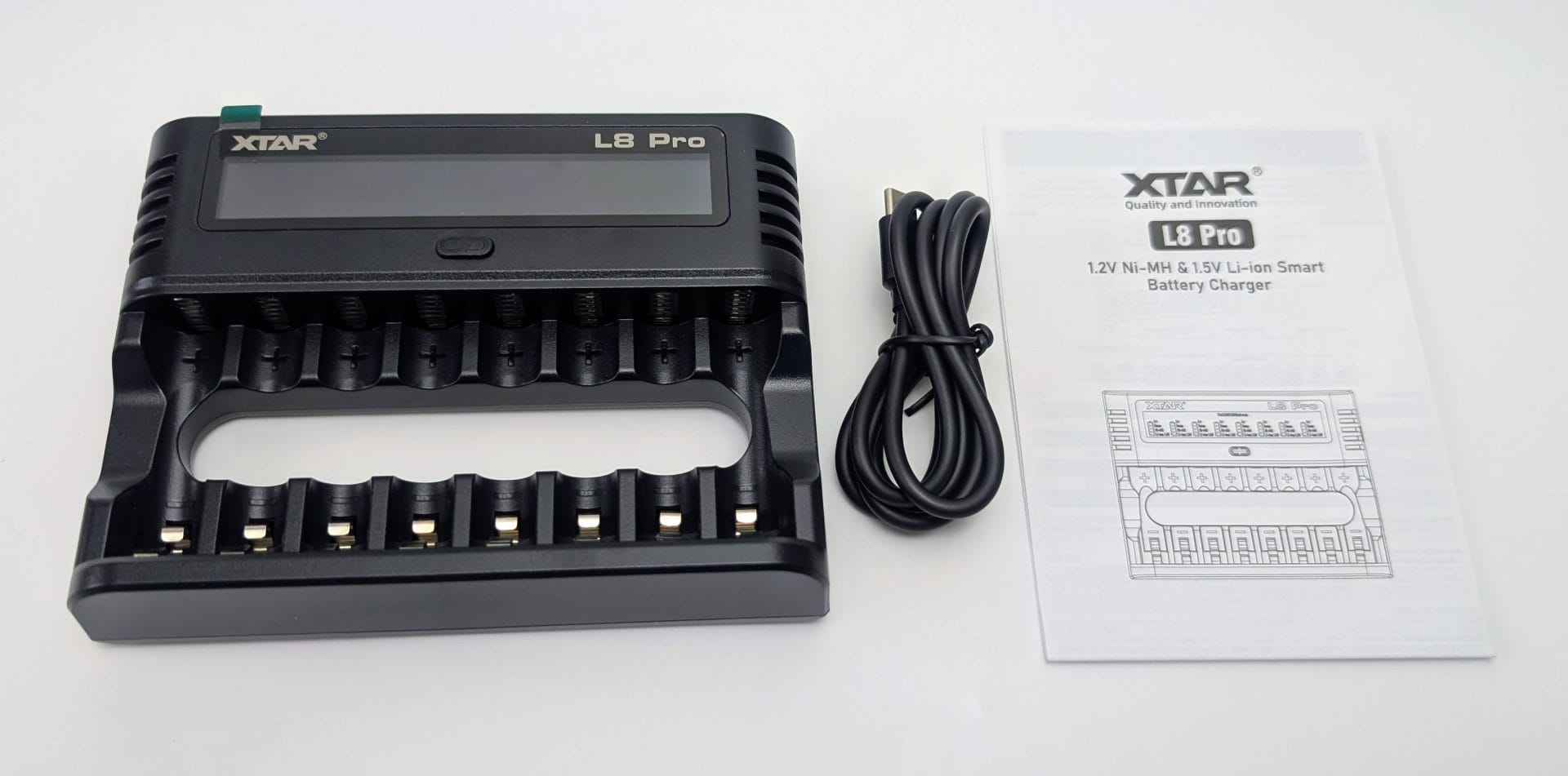 Black XTAR L8 Pro smart battery charger shown from the front, featuring an LCD display, eight AA/AAA battery slots with spring contacts and polarity markings, and a large finger-grip cutout. To the right are a coiled black USB cable and the white instruction manual titled “XTAR L8 Pro 1.2V Ni-MH & 1.5V Li-ion Smart Battery Charger.”