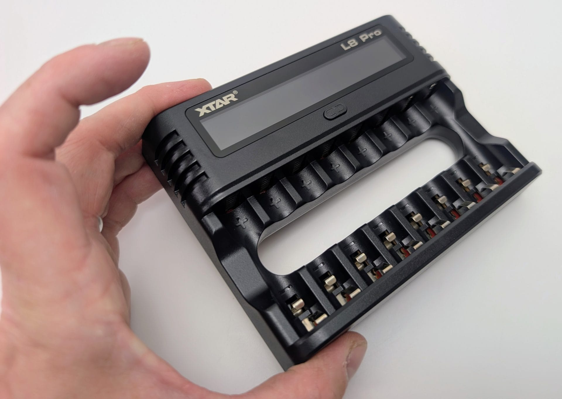 Close-up of a hand holding the black XTAR L8 Pro smart battery charger, angled to show the blank LCD screen, “XTAR” and “L8 Pro” branding, eight AA/AAA battery slots with positive polarity markings and metal spring contacts, and the large central finger-grip cutout.