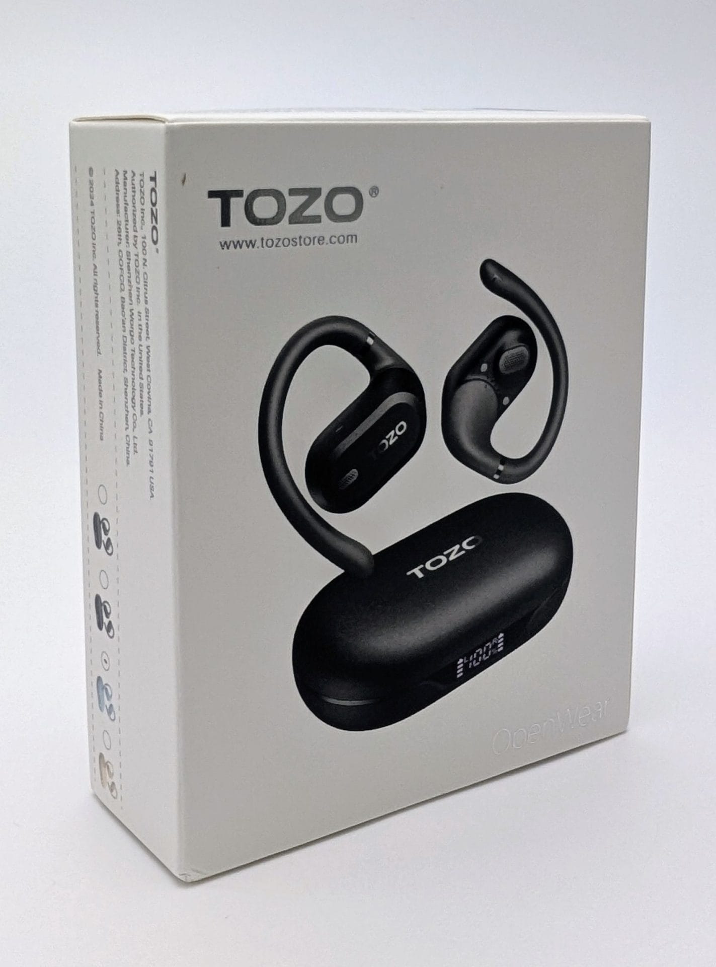 White product packaging box for TOZO OpenWear open-ear wireless earbuds. The front of the box shows black hook-style earbuds and a matching black charging case with a small digital display. The box is photographed at an angle against a plain white background, with the TOZO logo, website, and “OpenWear” branding visible.
