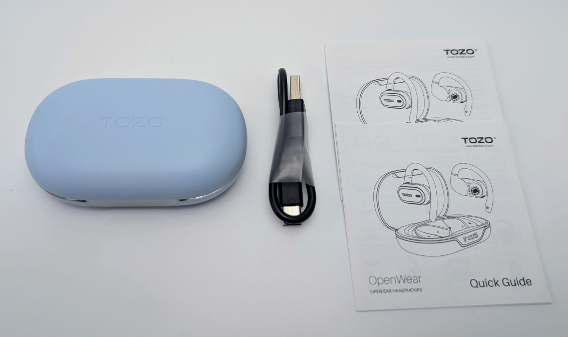 Light blue TOZO OpenWear charging case, a folded black USB charging cable, and two white Quick Guide booklets with line drawings of the open-ear headphones, arranged on a plain white surface.