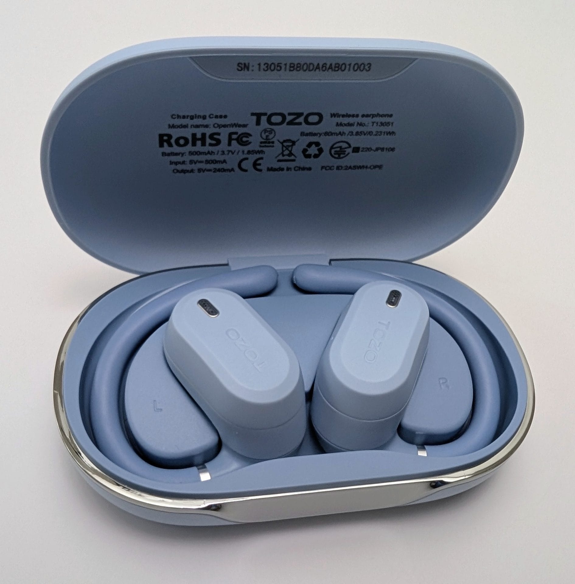 Open light blue TOZO OpenWear charging case with both open-ear earbuds docked inside, showing L and R markings, TOZO logos on the buds, and product specifications printed on the inner lid.
