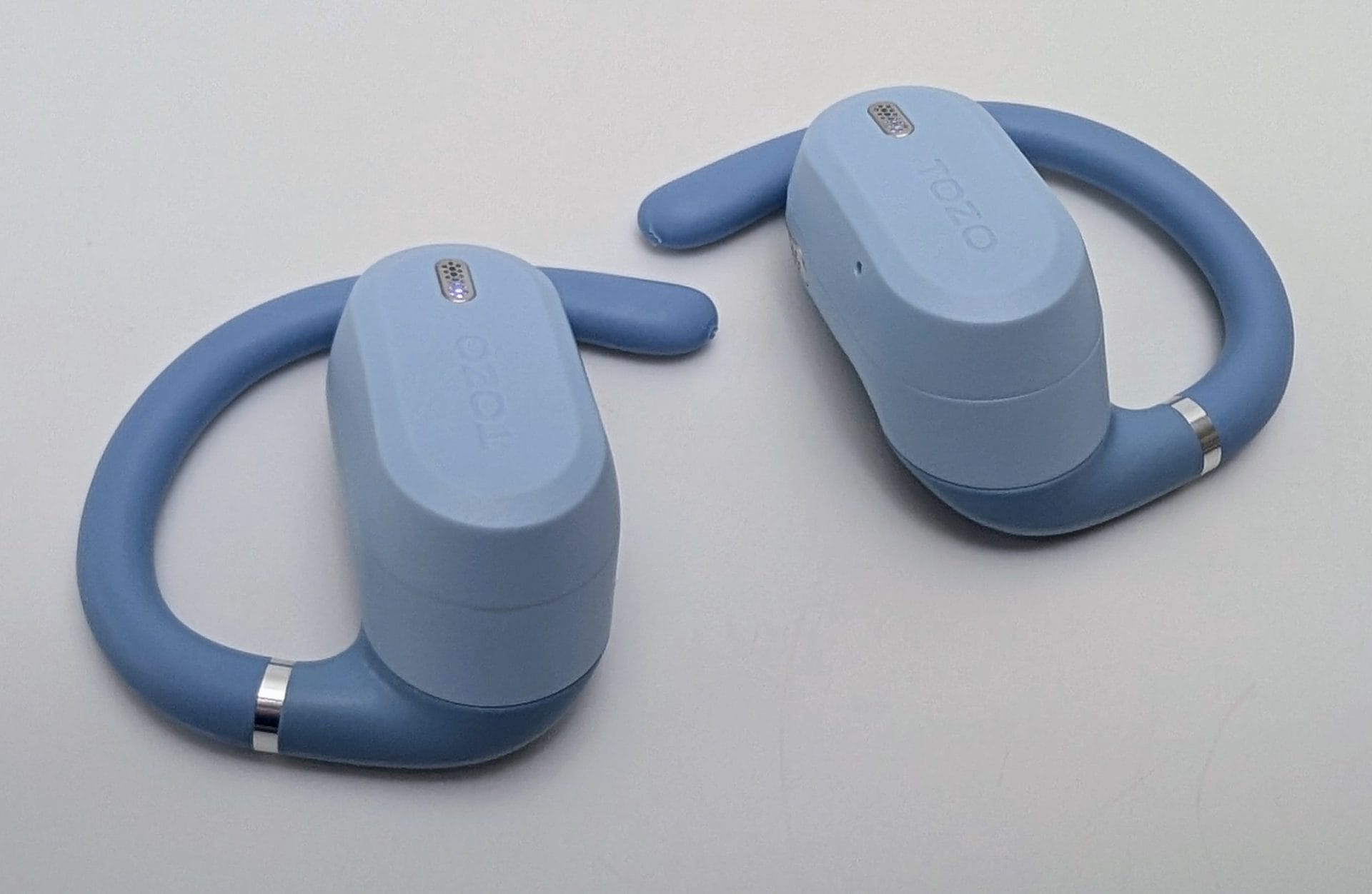 Pair of light blue TOZO OpenWear open-ear earbuds lying on a white surface, showing the flexible hooks with silver rings, TOZO logos, and charging contacts on top.
