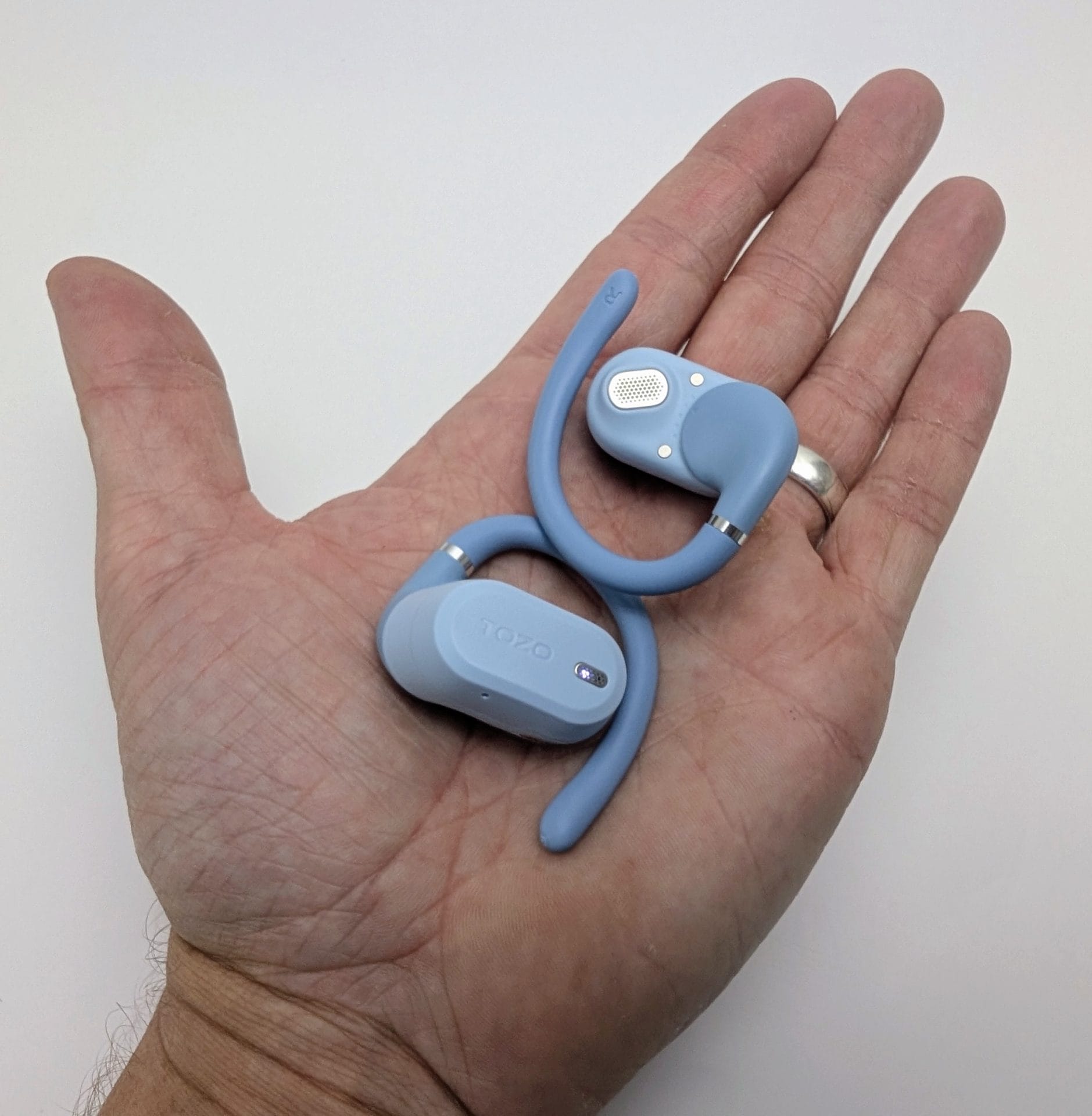 A person’s open left hand holding a pair of light blue TOZO OpenWear open-ear earbuds with flexible hooks, showing the speaker grille on one bud and the TOZO logo on the other, against a white background.