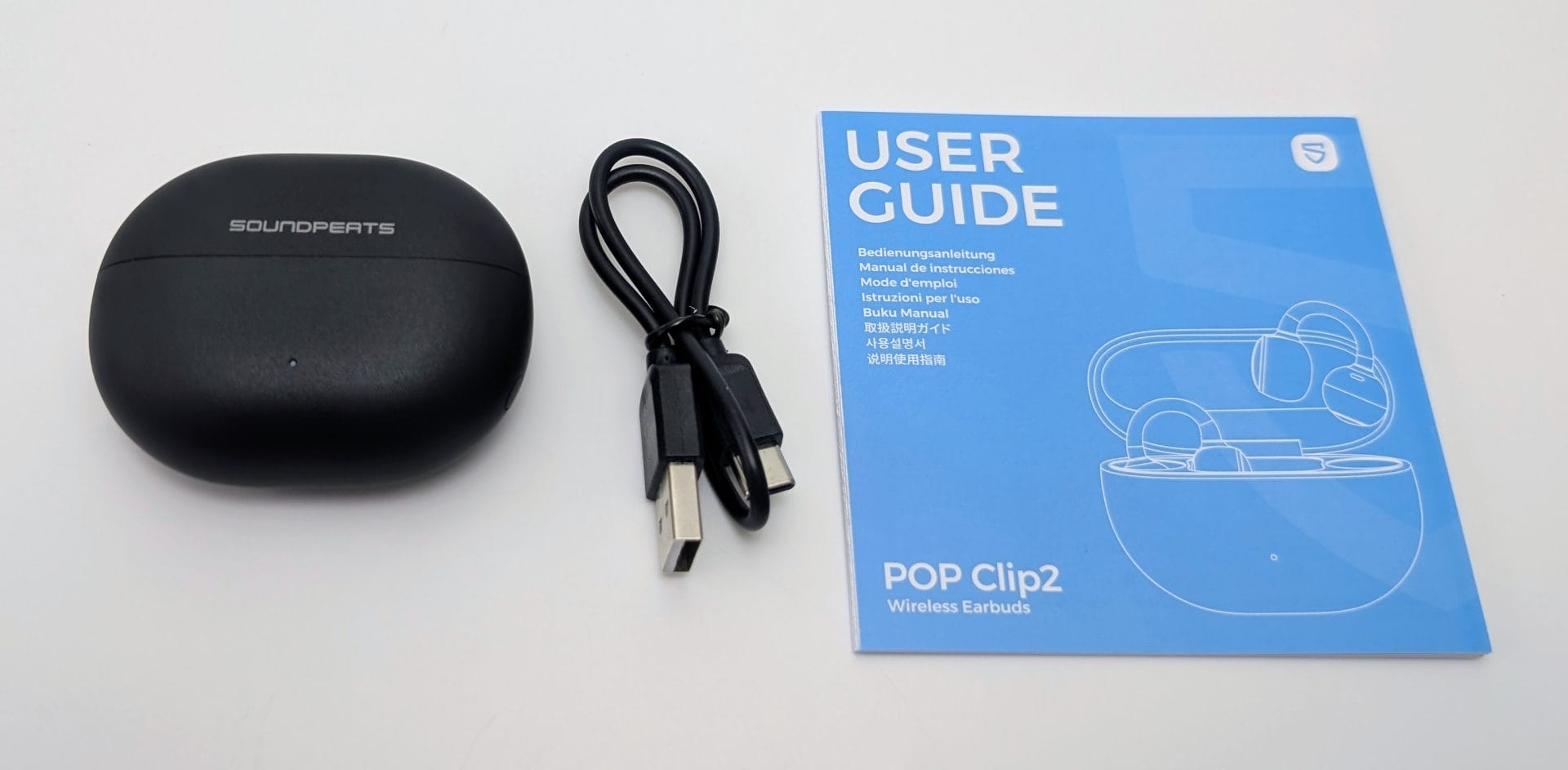 SoundPEATS UU2 Unboxed SoundPEATS UU2 (POP Clip2) accessories on a white surface: a matte black oval charging case with the SOUNDPEATS logo, a coiled black USB-A to USB-C cable, and a blue multilingual user guide titled “USER GUIDE” and “POP Clip2 Wireless Earbuds,” featuring a line drawing of the open case with clip-on earbuds inside.