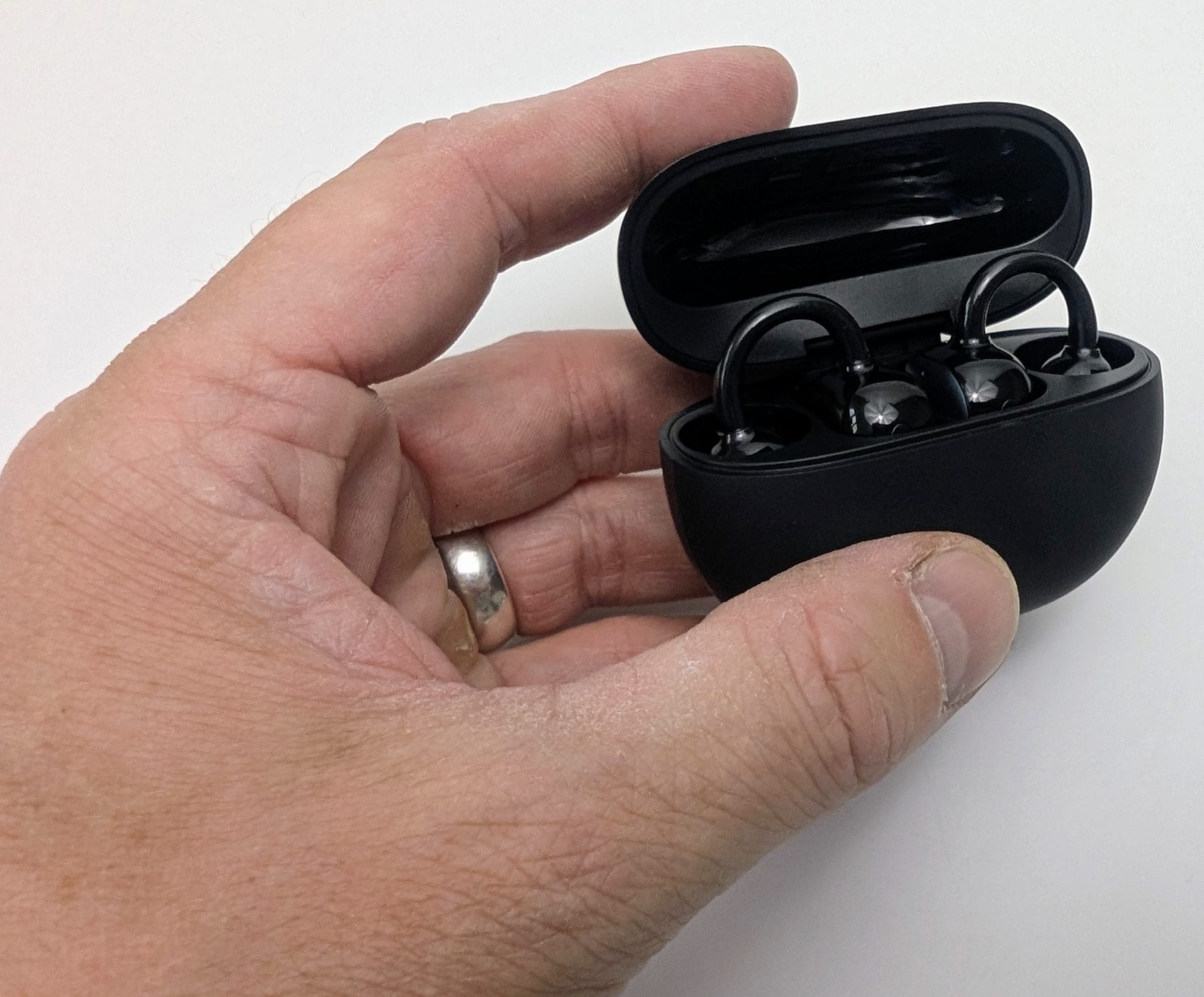 SoundPEATS UU2 A left hand wearing a silver ring holds an open matte-black SoundPEATS UU2 (POP Clip2) charging case against a white background. The lid is flipped up, revealing two glossy black clip-on earbuds seated in the case, with their U-shaped hooks visible.