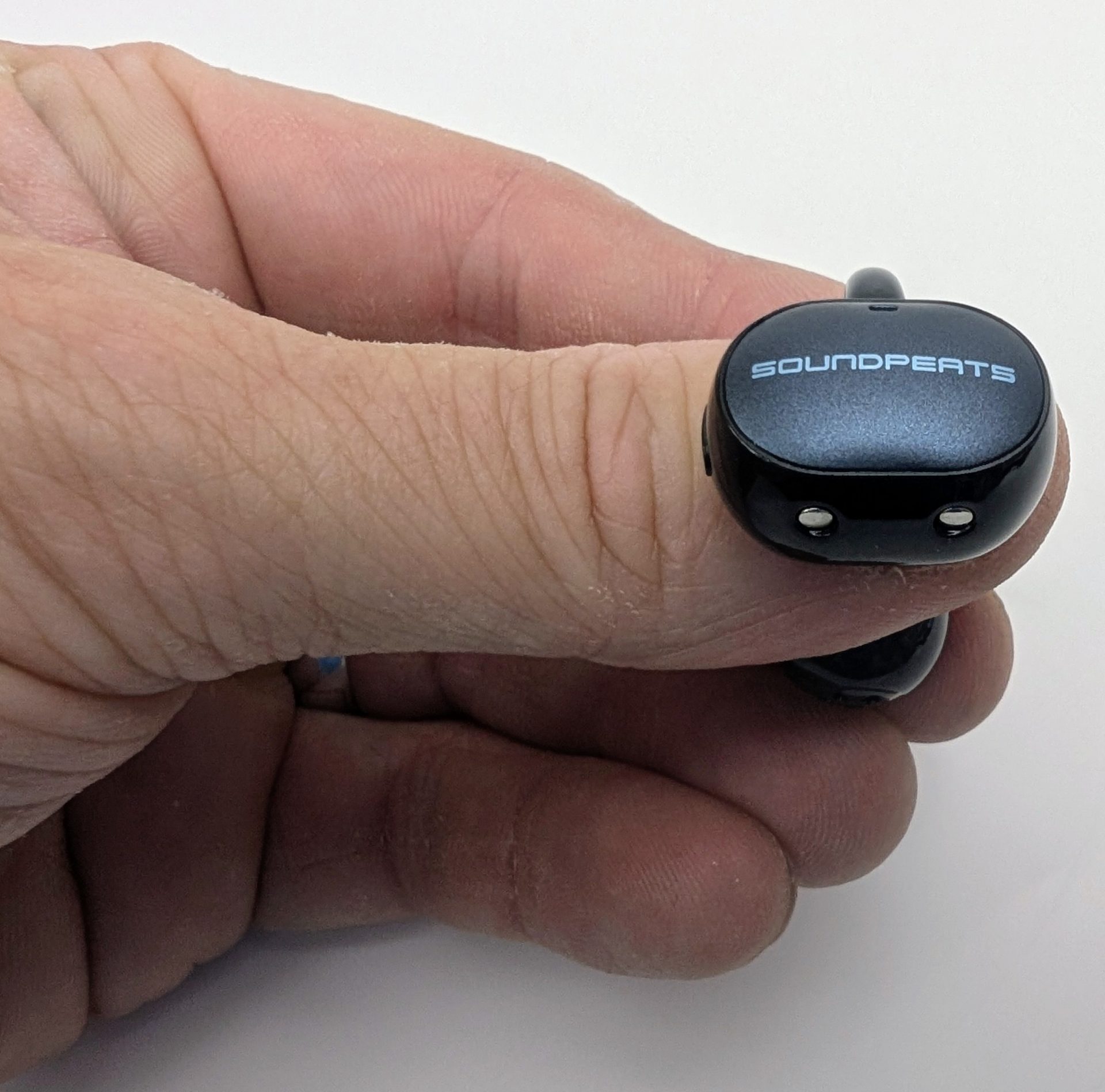 SoundPEATS UU2 Close-up of a hand holding one black SoundPEATS UU2 (POP Clip2) clip-on earbud. The inner face of the control body is visible, printed with “SOUNDPEATS” in light blue and two small gold charging contacts along the bottom edge. The U-shaped hook and spherical speaker housing extend behind the thumb.