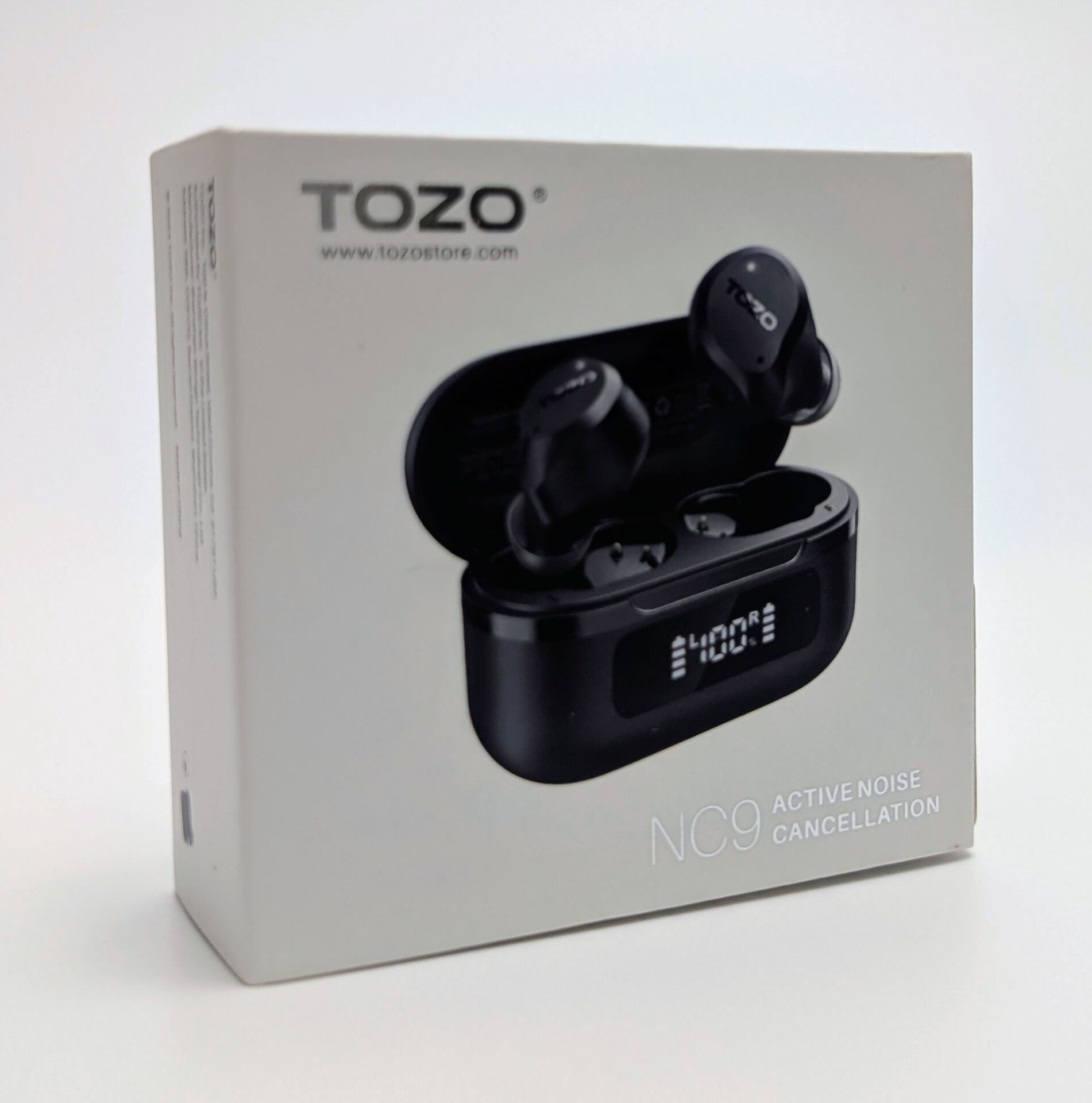 TOZO NC9 Earbuds White product box for the TOZO NC9 Active Noise Cancellation earbuds, photographed at an angle on a plain white background. The front shows the black TOZO logo and website, a large image of the black charging case with lid open and one earbud resting outside it (both marked TOZO), a digital display reading 400 on the case, and the text “NC9 ACTIVE NOISE CANCELLATION” at the bottom.
