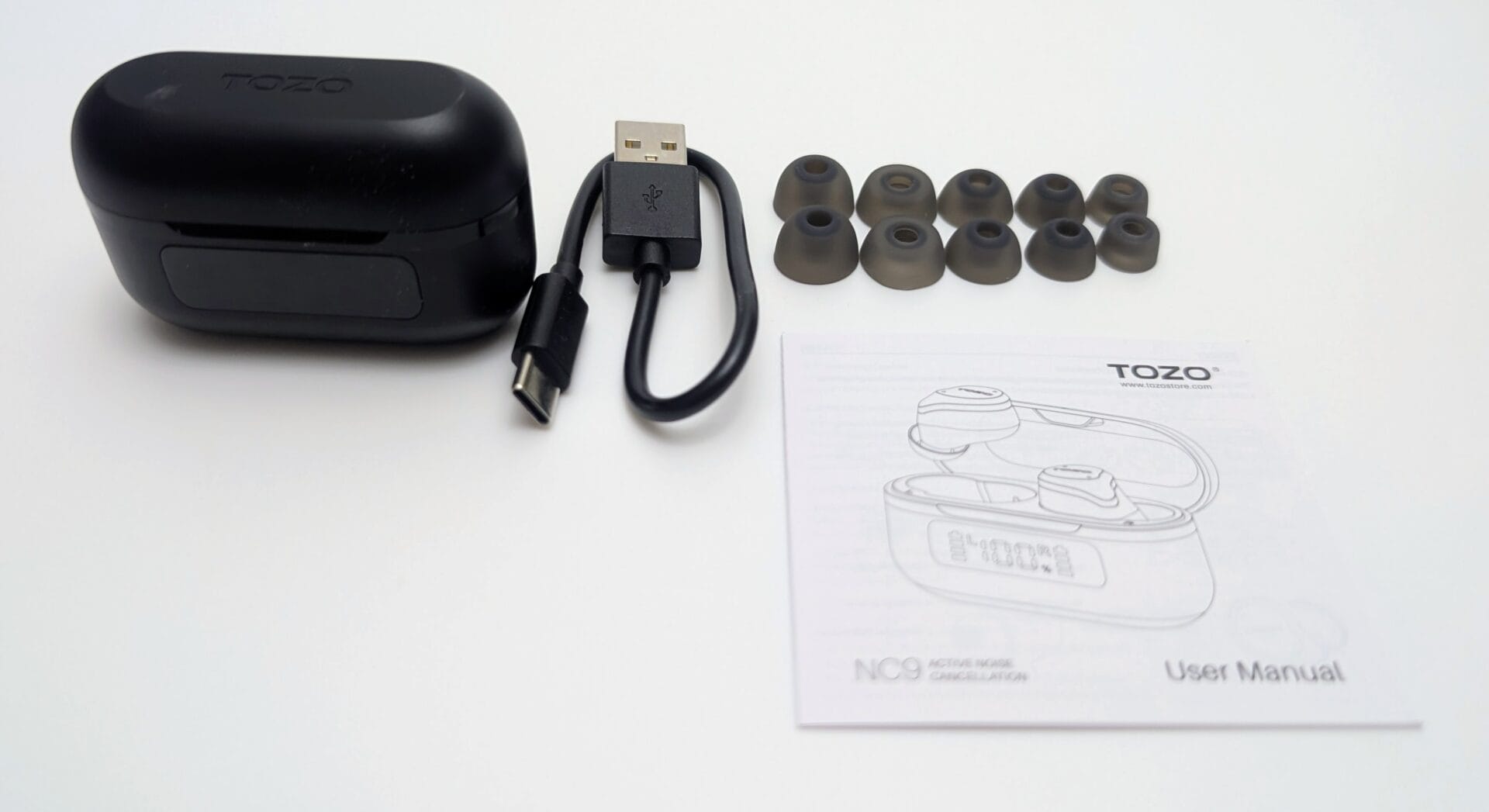 TOZO NC9 Earbuds Contents of the TOZO NC9 Active Noise Cancellation earbuds laid out on a white surface: a closed black charging case with TOZO logo on the lid, a black USB-A to USB-C charging cable, a set of translucent gray silicone ear tips in multiple sizes, and a white user manual featuring a line drawing of the open case, TOZO branding, and the text “NC9 ACTIVE NOISE CANCELLATION User Manual.”