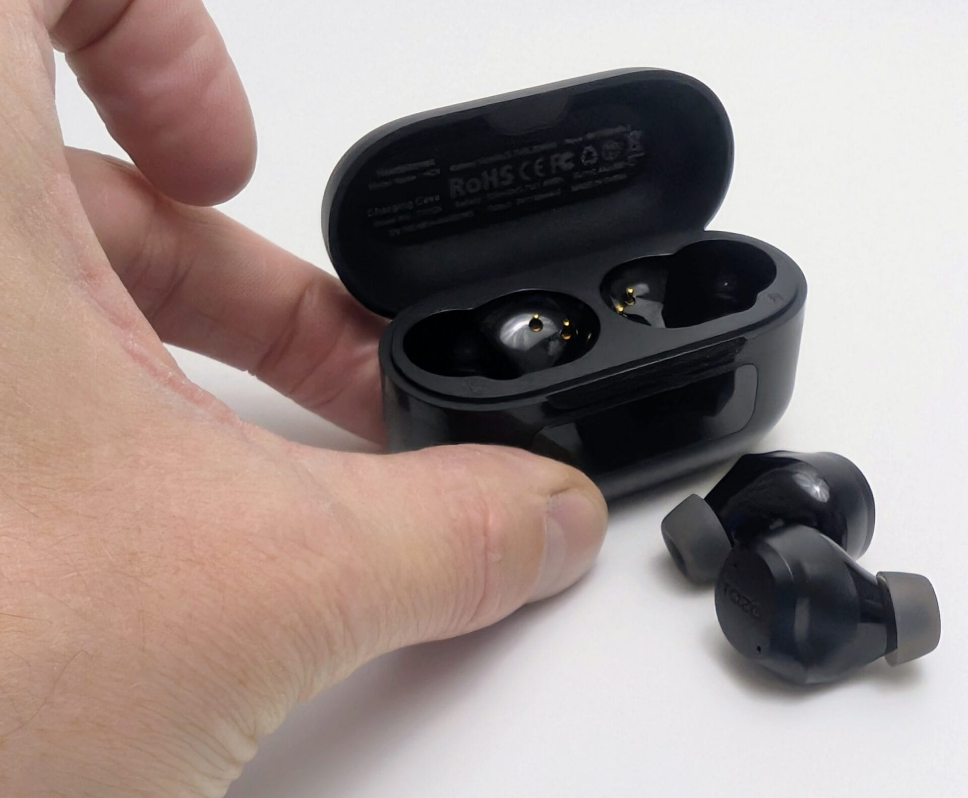 TOZO NC9 Earbuds A hand holds open a black TOZO NC9 charging case revealing one earbud seated in its slot with gold charging contacts visible, while the second earbud with a gray silicone tip rests outside the case; regulatory markings including RoHS and CE appear on the inside of the lid, set against a white background.