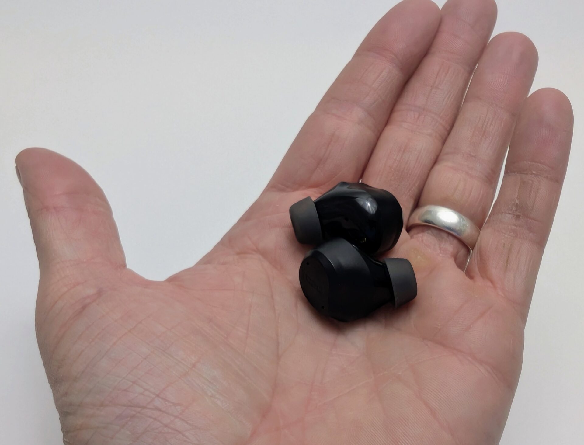 TOZO NC9 Earbuds Two black TOZO NC9 wireless earbuds resting in an open palm, one fitted with a dark silicone ear tip and both showing a glossy finish with the TOZO logo visible on one, next to a silver ring on the ring finger, against a plain white background.