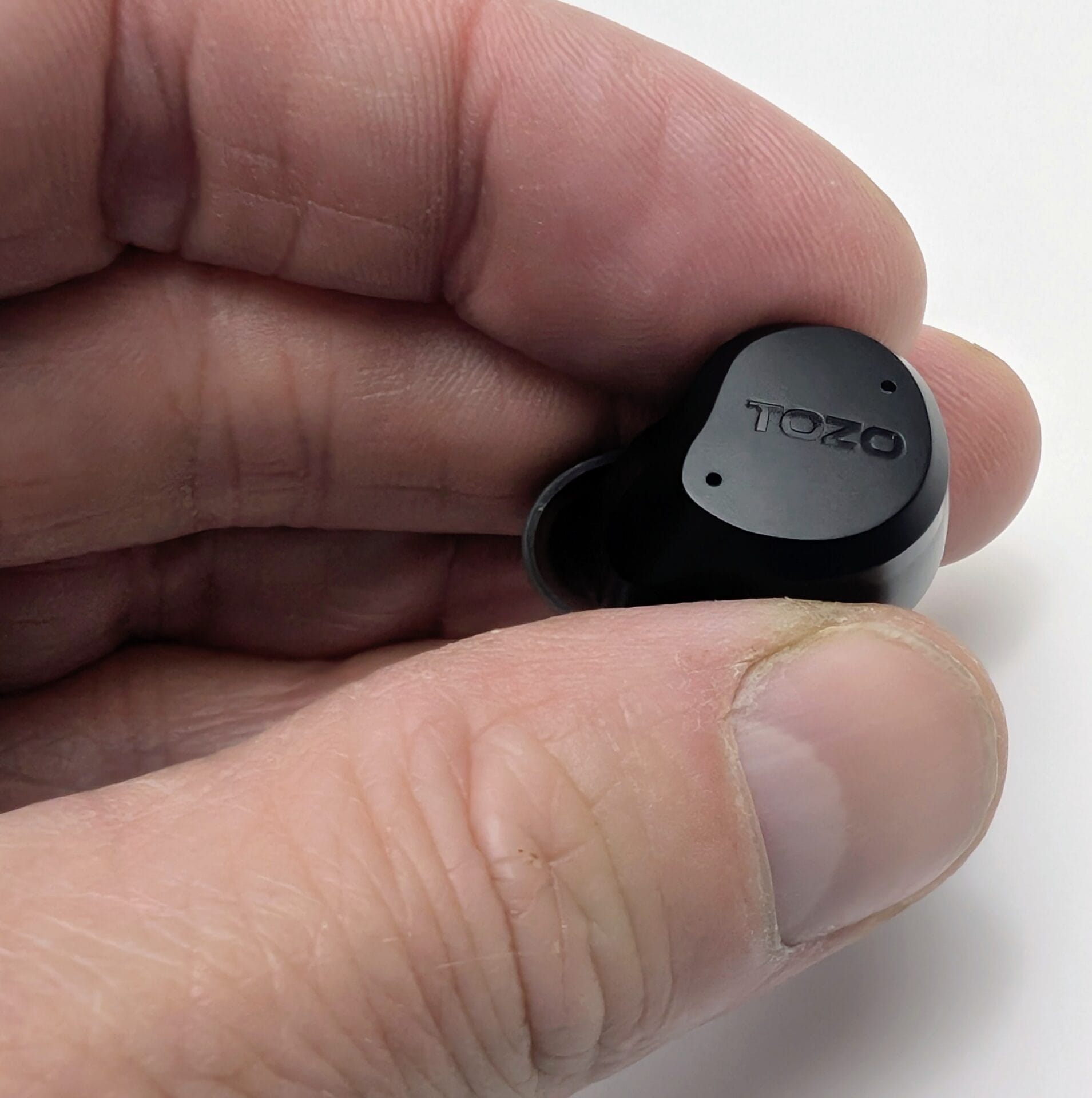 TOZO NC9 Earbuds Close-up of a hand holding a single black TOZO NC9 wireless earbud, showing the outer face with the embossed TOZO logo and two small microphone holes, against a plain white background.